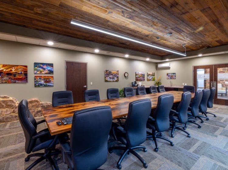 A modern conference room with a large wooden table and leather chairs. The space features a rustic wooden ceiling, stone accents, and vibrant artwork on the walls.