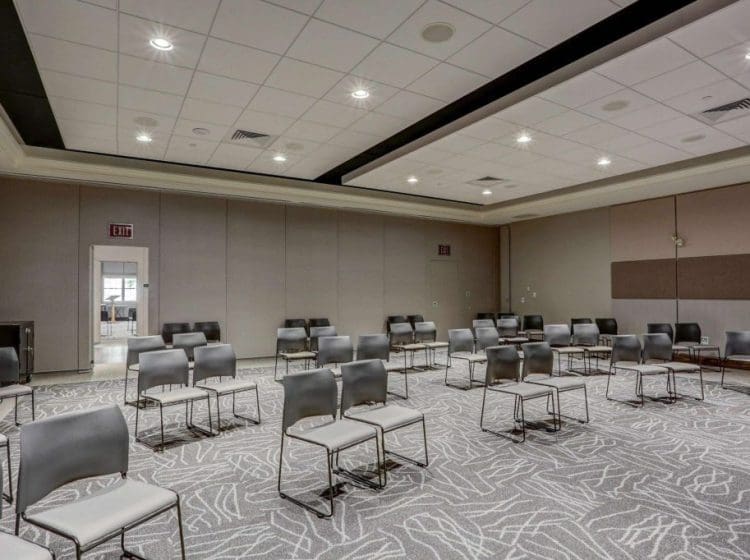 Meeting room with gray walls, modern carpet flooring, and spaced-out seating. The ceiling has recessed lighting, and an exit door is visible at the back
