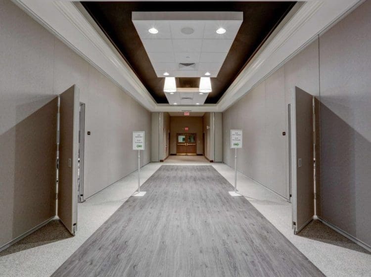 Spacious hallway with light wood flooring and neutral wall colors. The ceiling features recessed lights and modern fixtures, leading to another room at the end.