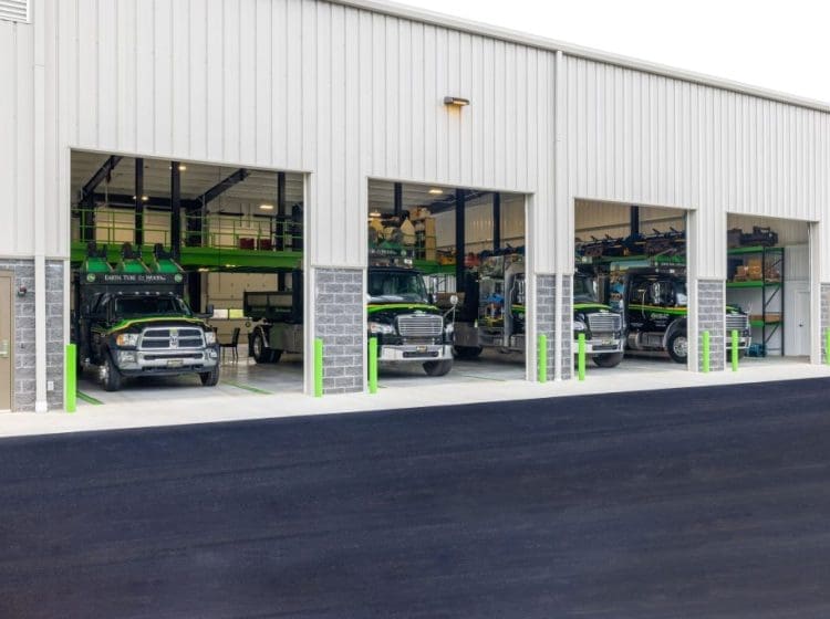 A truck maintenance bay with multiple large vehicles parked inside a modern facility. The exterior of the building features industrial design with metal siding and stone accents