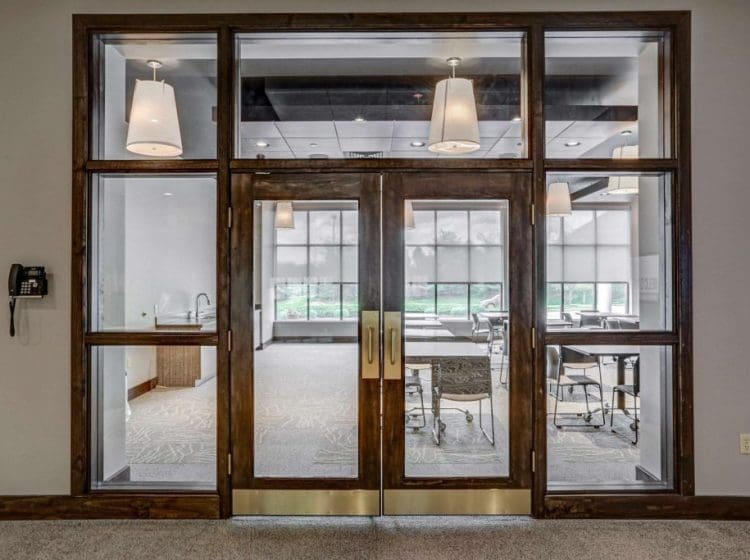 Entrance to a conference room with large glass double doors. The room is equipped with tables, chairs, and pendant lighting, with natural light coming through large windows