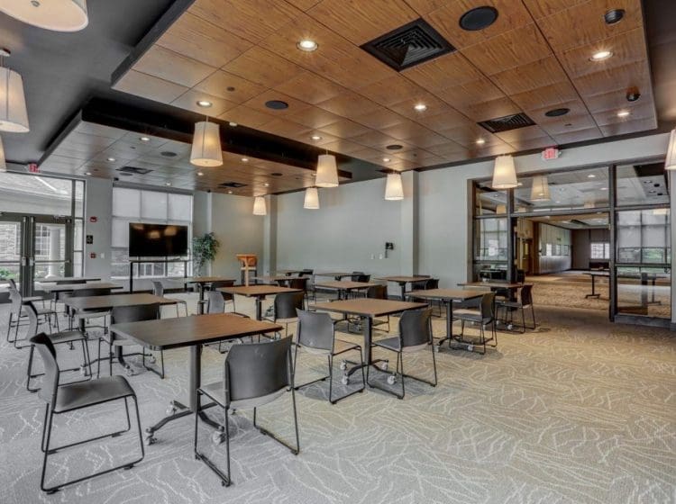 Conference room with wood-paneled ceiling and pendant lights. Tables and chairs are arranged throughout the room, with glass doors leading to an adjoining space.