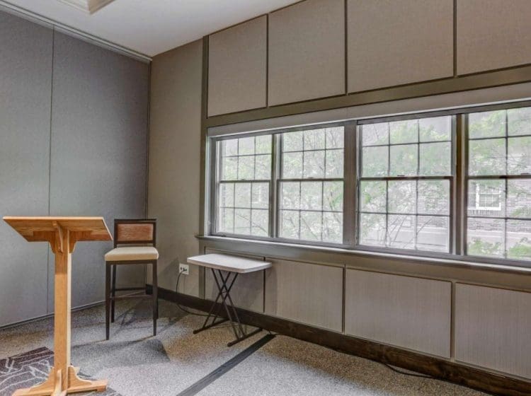 Small presentation area with a wooden podium, a chair, and large windows allowing natural light into the room. Neutral-colored walls and modern carpet complete the setup.