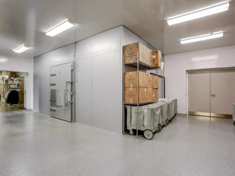 Industrial storage area with large metal racks holding boxes, carts, and supplies. The space has bright fluorescent lighting and polished floors for easy cleaning