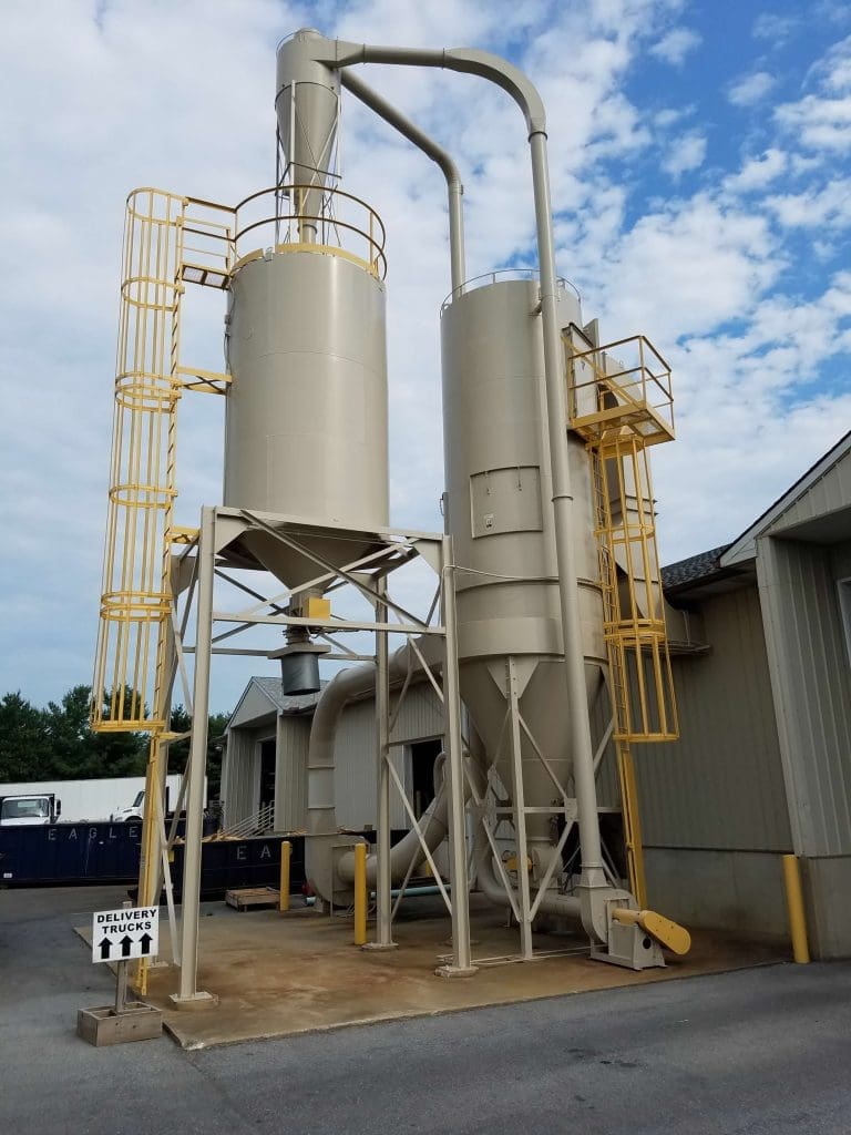 Two industrial silos with visible piping, yellow ladders, and maintenance equipment nearby.