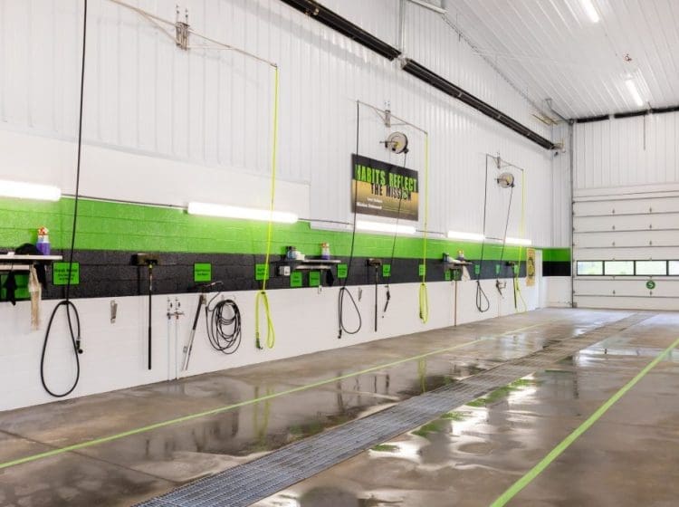 Interior view of a commercial vehicle wash bay featuring bright white walls with green and black accent stripes. The space includes multiple washing stations with hoses, cleaning tools, and equipment hanging on the wall. The floor is clean and wet, reflecting the overhead lights