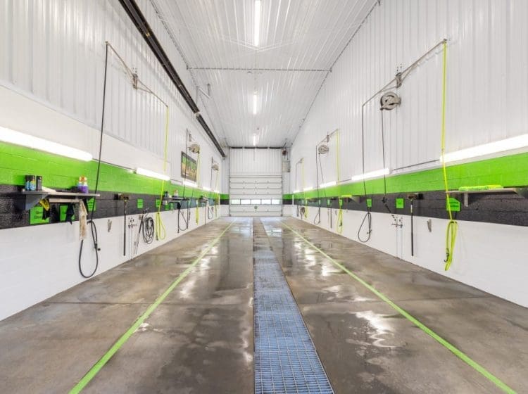 An interior view of a clean commercial vehicle wash bay featuring bright white walls and green accents. The facility includes multiple washing stations and overhead hoses