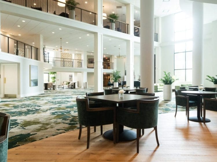 Spacious, elegant lobby with high ceilings, large windows, and green upholstered chairs. The room features a mix of modern and traditional design elements, with a mezzanine and ample seating