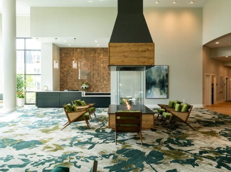 Modern lounge area with a central glass fireplace surrounded by mid-century style chairs. The space has patterned carpet and warm, ambient lighting