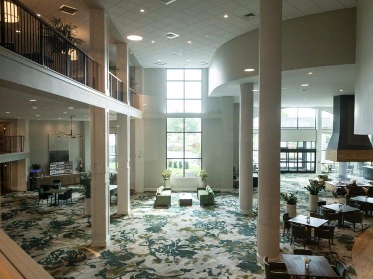 Bright and open atrium-style lobby with floor-to-ceiling windows, a mix of seating areas, and a large, central fireplace. The space is decorated with plants and natural light floods the room