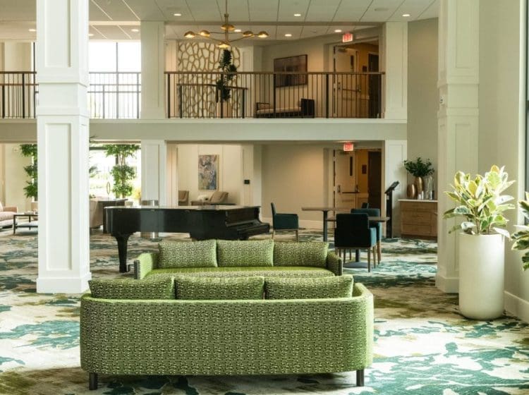 Lobby seating area with green upholstered sofas, a grand piano in the background, and high ceilings. The space features multiple levels with balconies and elegant decor.