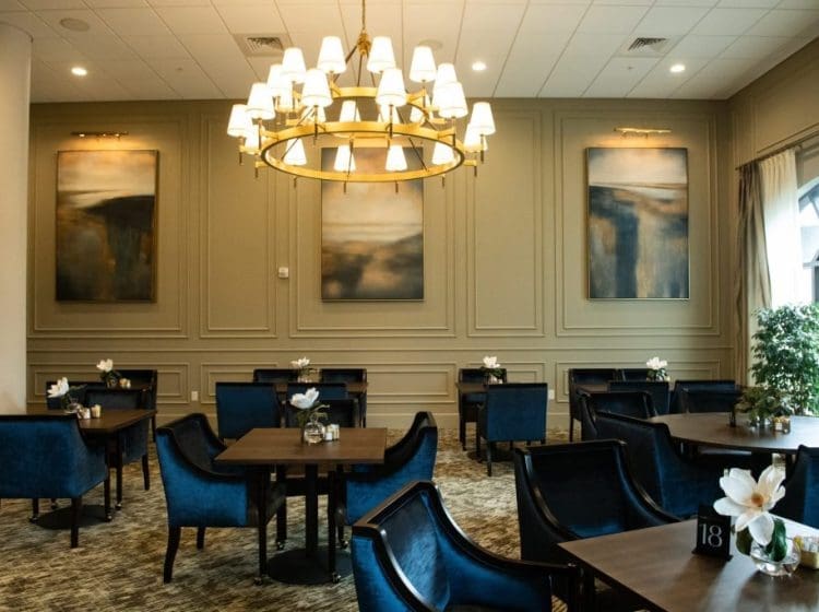 Cozy dining room with dark blue velvet chairs, wooden tables, and a large chandelier. Artwork and soft lighting create a refined, intimate atmosphere.