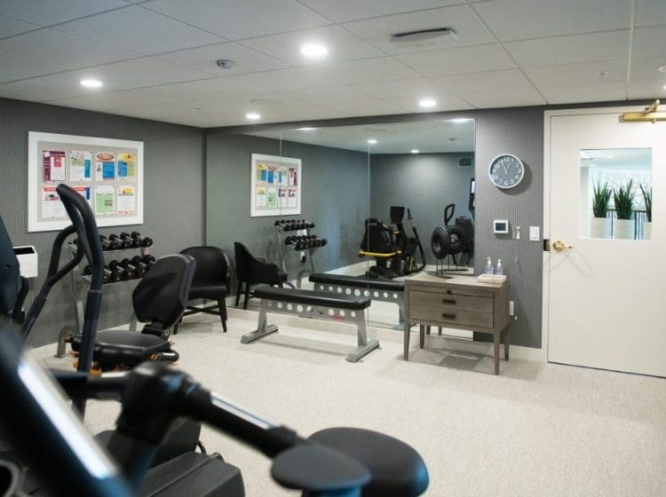 Small fitness room equipped with weights, exercise machines, and a mirrored wall. The room has a modern design with light gray tones and efficient use of space.