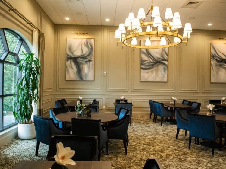 Elegant dining room featuring dark blue velvet chairs, round wooden tables, and a large, modern chandelier. Abstract artwork and large arched windows bring in natural light, complementing the luxurious ambiance.
