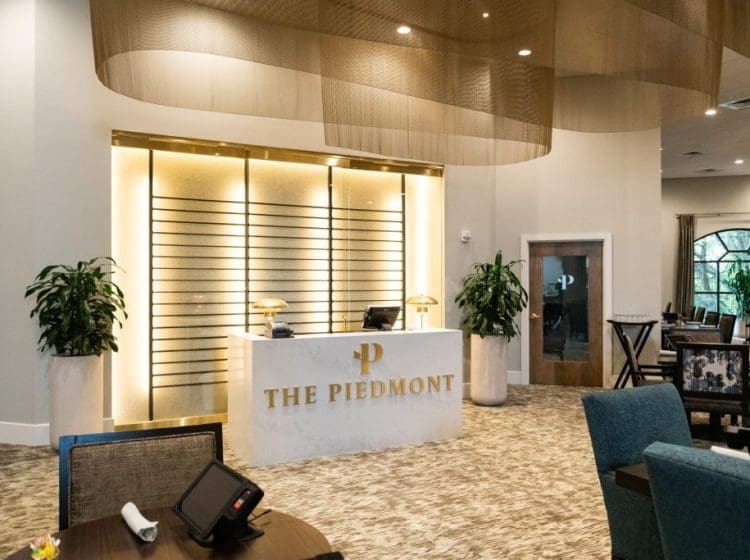 Sophisticated reception area with a marble desk labeled 'The Piedmont.' The background features a warm gold accent wall and modern lighting, with plants on either side of the desk