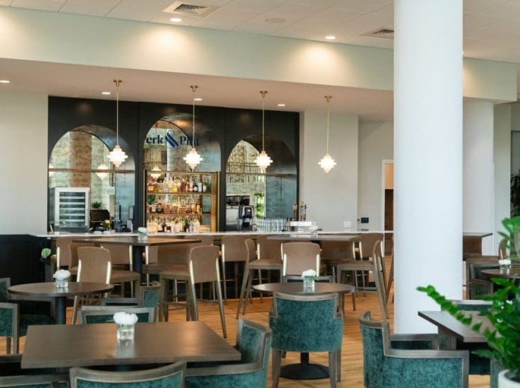 Stylish bar area with green upholstered chairs and pendant lights. The bar is lined with a variety of drinks and features a sophisticated, arched mirror backdrop.