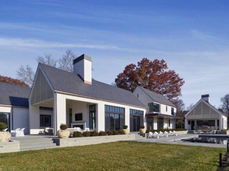 Modern farmhouse-style home with multiple gabled roofs, white exterior, and expansive windows. The property features an outdoor patio with seating, a pool, and a large lawn.
