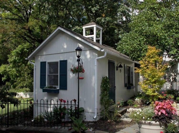 Small white garden shed with blue shutters and surrounded by a landscaped yard and a black fence.