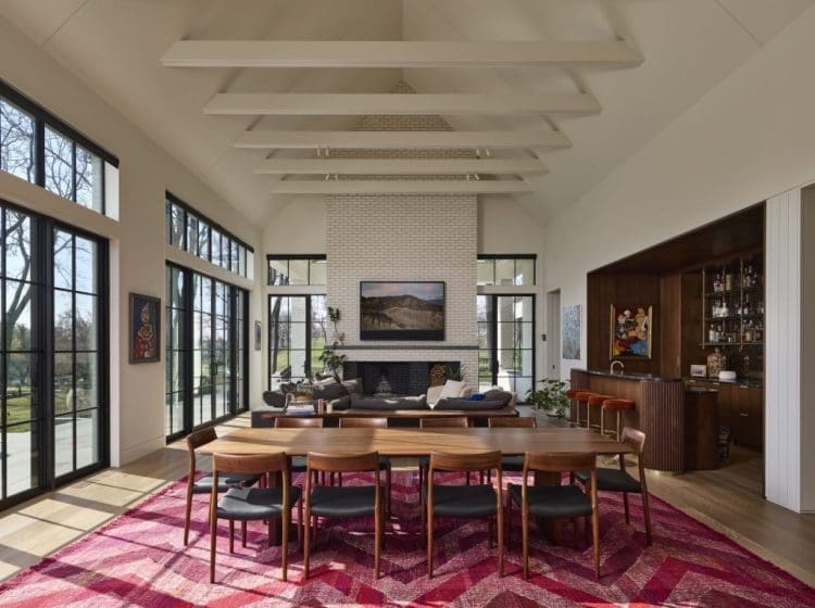 Large open living room with exposed beams, a white brick fireplace, and floor-to-ceiling windows. A wooden dining table is centered on a red-patterned rug, with modern décor throughout.
