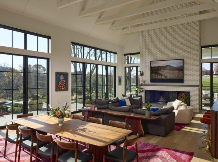Spacious living and dining room area with floor-to-ceiling windows, showcasing an outdoor view. The room features a large brick fireplace, modern furniture, and a wooden dining table.