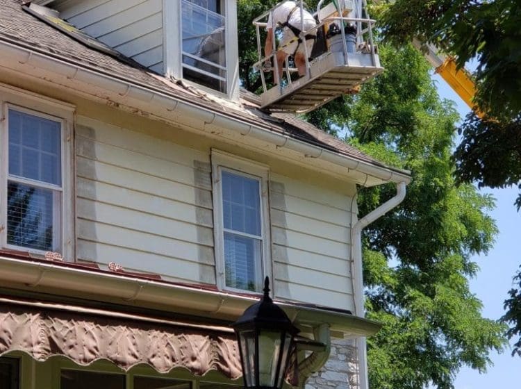 Worker on a lift painting the upper story of a house with siding and stone walls.