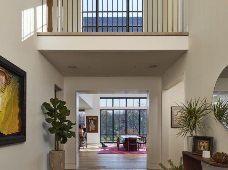 Bright entryway in a contemporary home with high ceilings and a second-floor balcony overlooking the foyer. The hallway leads to a dining area with large windows offering a view of the outdoors.