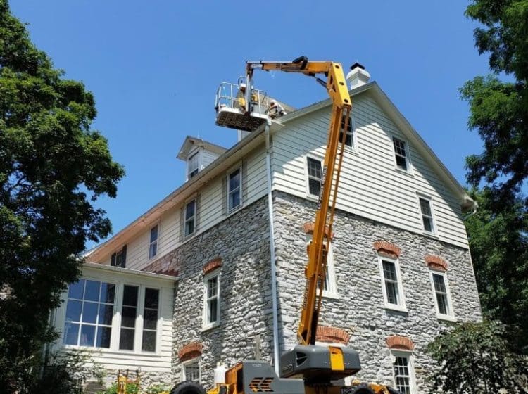 Yellow articulated lift used for exterior work on a three-story house with stone and siding.