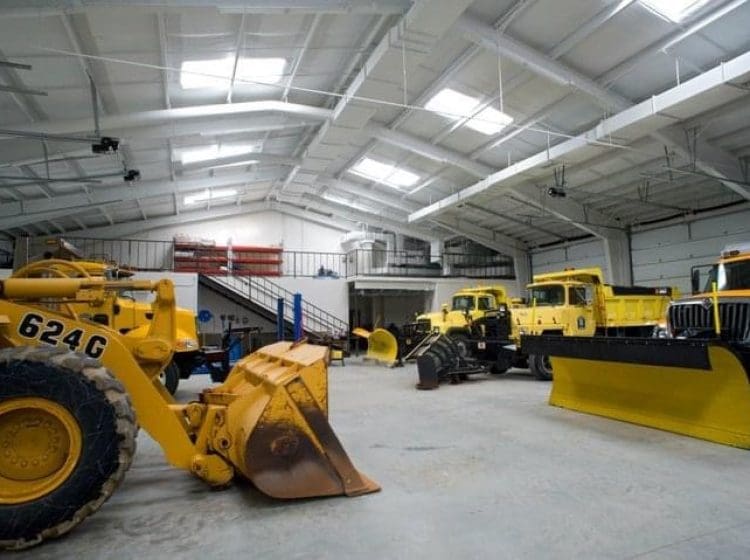 Large truck garage with various industrial vehicles and plows, including yellow trucks and equipment under bright overhead lights.