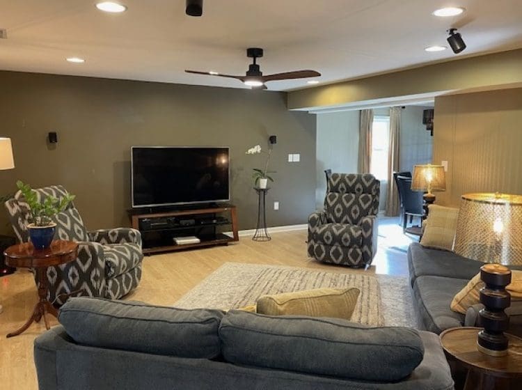 Spacious living room with olive-green walls and a mix of comfortable furniture, including a sectional sofa, patterned armchairs, and a large TV mounted on the wall. A ceiling fan with recessed lighting adds a modern touch.