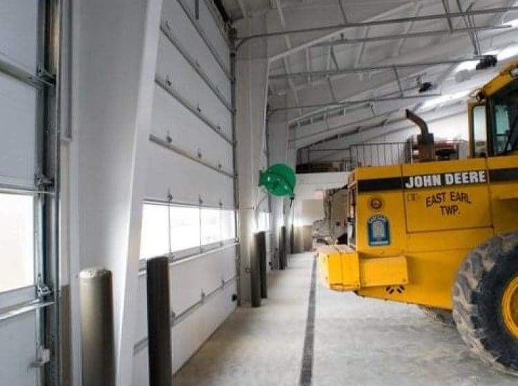 Interior of a truck garage with a yellow John Deere vehicle and large roll-up doors. The garage has high ceilings with bright lighting and a trench drain system.