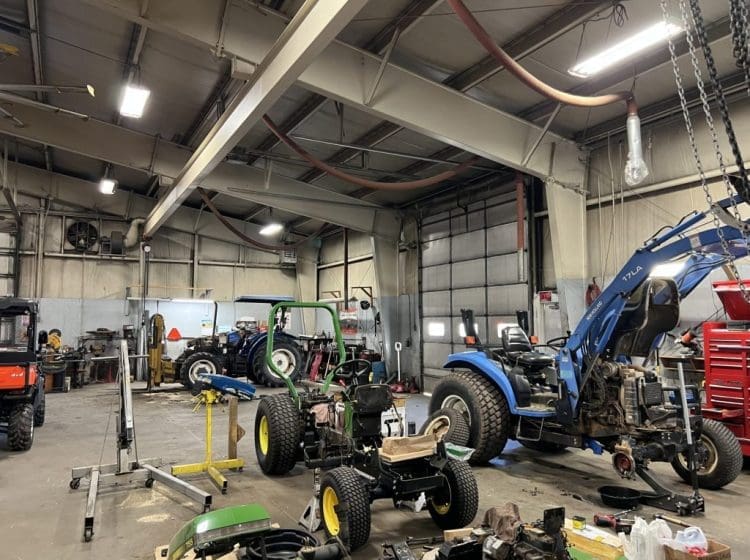 Industrial workshop featuring blue and green tractors, workbenches, and equipment in a metal building.