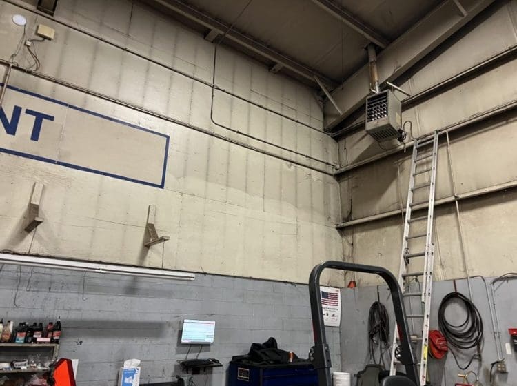 Corner of an industrial workshop showing a ladder, wall-mounted heater, and various tools and equipment.