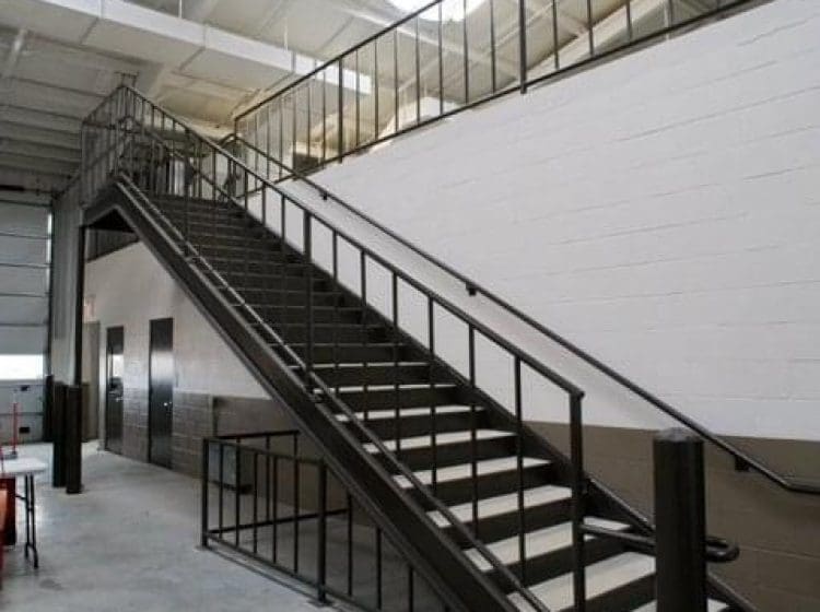 Metal staircase with black handrails leading up to a mezzanine in an industrial building. The walls are painted in two-tone colors, and the floor is bare concrete