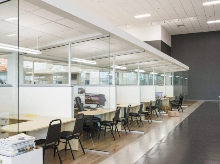 Modern office cubicles with glass walls, wood-look flooring, and computer workstations.