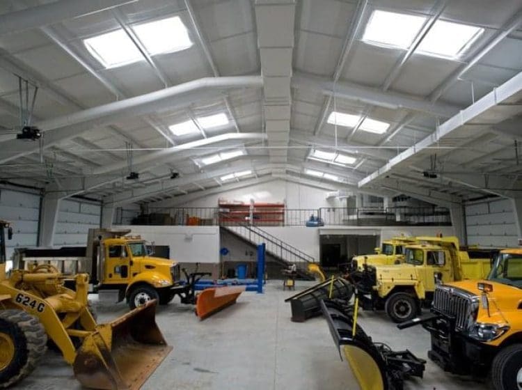 Interior of an industrial truck garage with bright overhead lighting, featuring several large trucks and plows stored in an organized fashion under the high ceilings