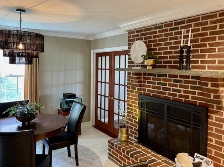 Close-up of a brick fireplace with a wooden mantle, decorated with candles and small decorative items. A modern black chandelier hangs above a round dining table with leather chairs, giving the room a cozy and stylish feel.