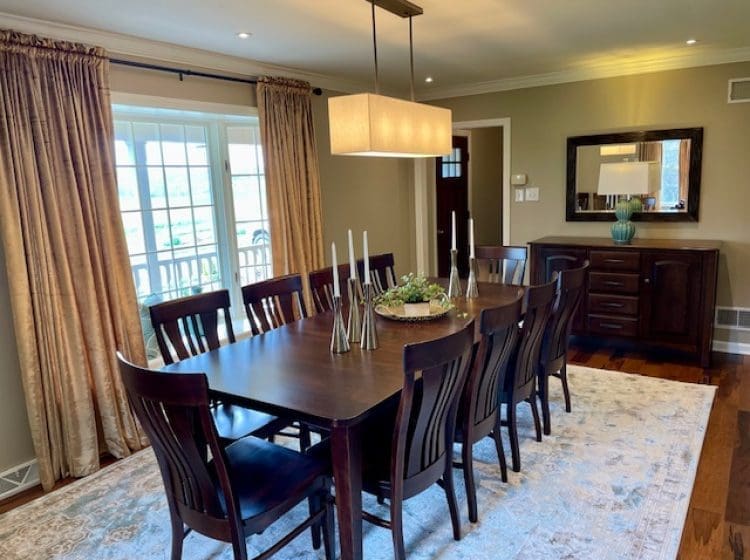 Elegant dining room with a large wooden table and eight matching chairs. Floor-length curtains frame a set of windows that allow natural light to fill the space. A soft rug and modern chandelier add sophistication to the room.