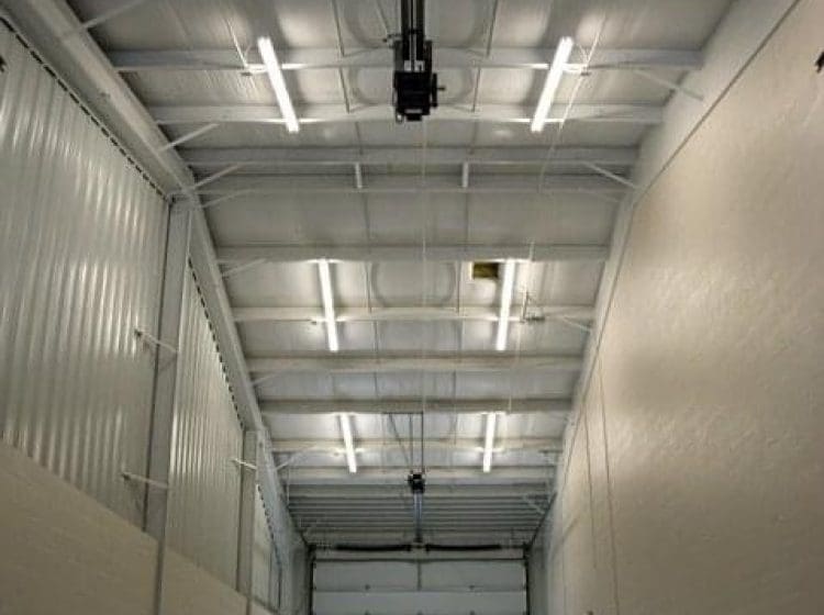 Long, narrow truck bay with roll-up doors at both ends, illuminated by fluorescent lighting and featuring a two-tone paint job on the walls.