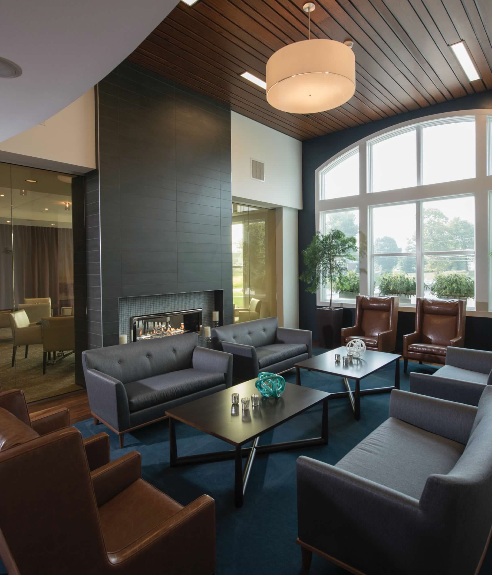 A modern office lounge with sleek furniture, a fireplace, wood-paneled ceiling, and large windows.