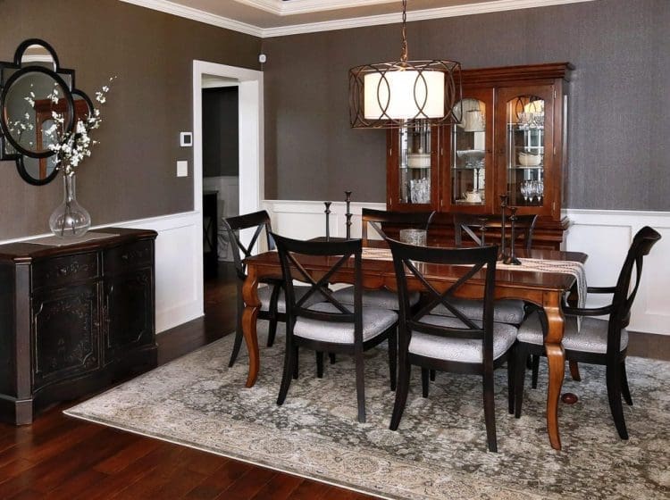A traditional dining room with dark wood furniture, gray walls, white wainscoting, and a patterned area rug.