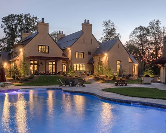 A large home with stucco exterior and modern design, illuminated by outdoor lighting, near a swimming pool at dusk.