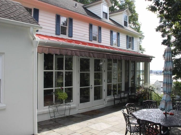 A colonial-style home with a red metal roof, large windows, and a patio with outdoor seating and landscaping.