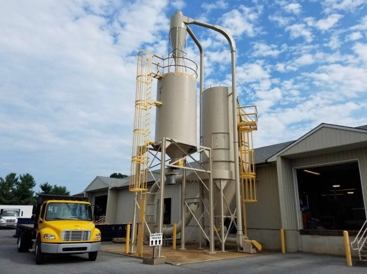Tall industrial silos with yellow safety ladders and platforms next to a tan building and a yellow delivery truck.
