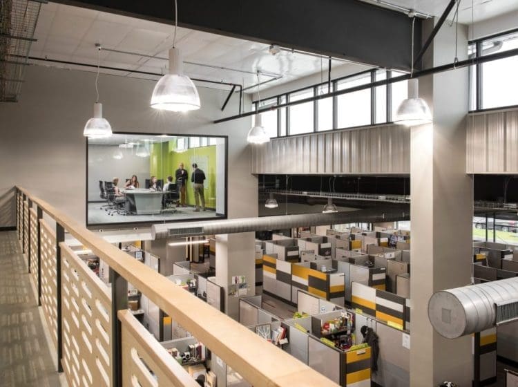 A modern office workspace with cubicles, a glass-walled conference room, and industrial-style lighting.