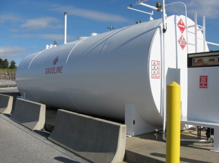 Large white gasoline storage tank with safety signage and yellow bollards in an outdoor industrial setting.