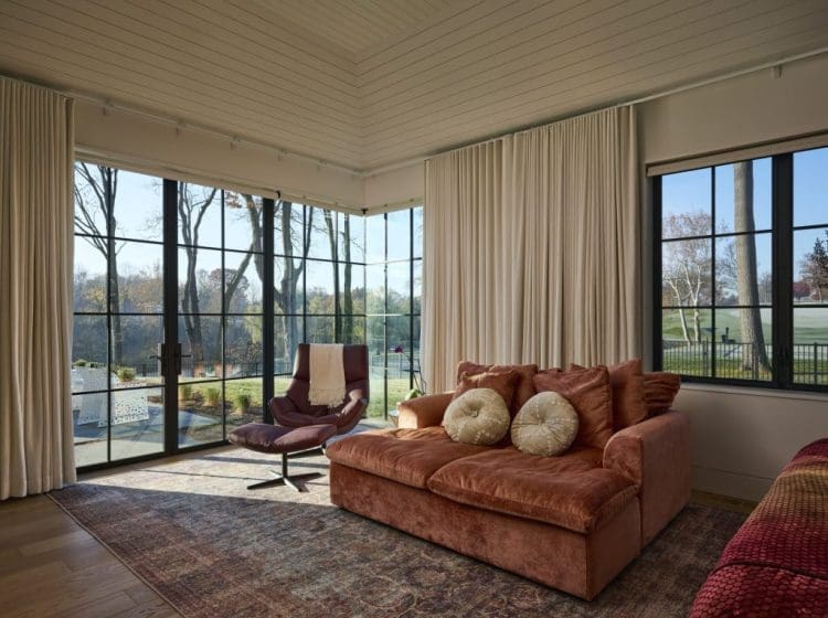 A sunroom with floor-to-ceiling windows, cream curtains, and modern furniture overlooking an outdoor garden.